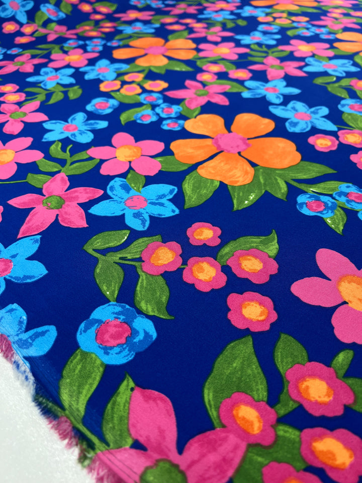 A vibrant polyester fabric with a blue backdrop is adorned with a floral pattern featuring pink, orange, and blue flowers, accompanied by green leaves. The design is colorful and lively, creating an eye-catching and cheerful motif perfect for designer fabrics like the Deluxe Print - Nursery - Nautical - 155cm from Super Cheap Fabrics.