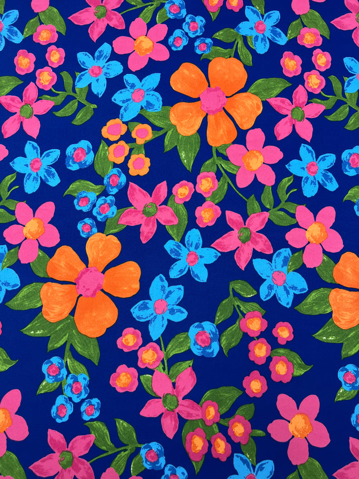 A vibrant pattern featuring colorful flowers in various sizes and colors, including orange, pink, and blue. The background is a rich blue color, which makes the flowers stand out prominently. Ideal for designer fabrics, Deluxe Print - Nursery - Nautical - 155cm by Super Cheap Fabrics features flowers interspersed with green leaves and buds for added elegance.