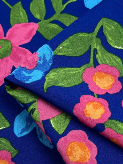 Close-up shot of a vibrant polyester fabric with a blue background featuring colorful floral patterns. The flowers are in shades of pink and orange, with green leaves and occasional blue accents. The Super Cheap Fabrics Deluxe Print - Nursery - Nautical - 155cm is slightly folded in the image, showcasing its versatile appeal.