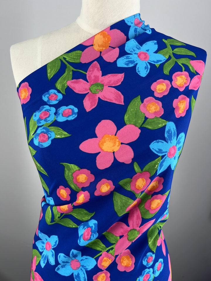 A mannequin draped with a brightly colored floral Deluxe Print - Nursery - Nautical - 155cm by Super Cheap Fabrics. The designer fabric features large, vibrant pink, orange, and blue flowers with green leaves on a deep blue background. The design is lively and energetic, with the flowers appearing in various sizes and orientations.