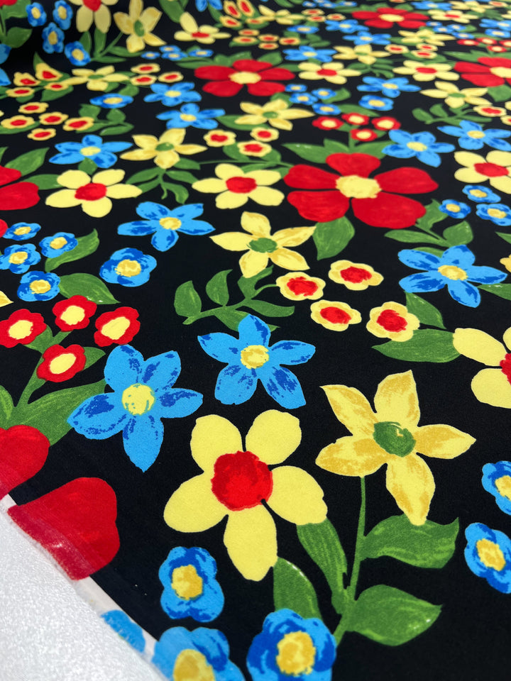 A Super Cheap Fabrics Deluxe Print - Nursery - Black - 155cm features an array of colorful flowers in red, yellow, blue, and green hues set against a black background. This designer fabric showcases flowers in various stages of bloom, creating a lively and cheerful design that's as versatile as it is stunning.
