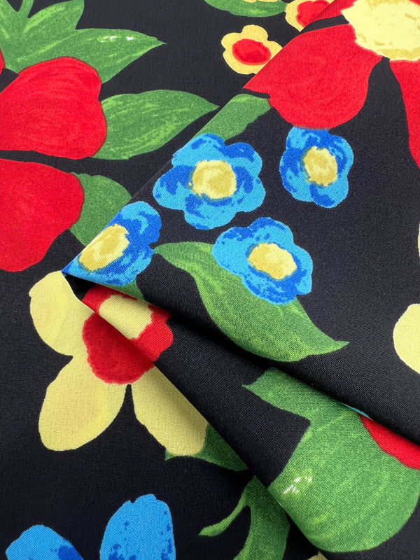 Close-up of designer fabric with a vibrant floral pattern, featuring large red, blue, yellow, and green flowers on a black background. The fabric is slightly folded, showcasing the colorful design in detail. Super Cheap Fabrics' Deluxe Print - Nursery - Black - 155cm is easy to care for and adds elegance to any project.