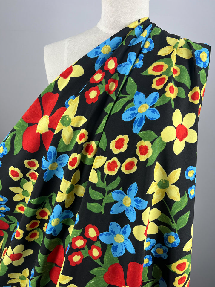 A person is partially visible, draped in a designer fabric adorned with vibrant red, yellow, and blue flowers and green leaves on a black background. Covering their upper body, the Deluxe Print - Nursery - Black - 155cm from Super Cheap Fabrics is arranged to showcase its stunning pattern.