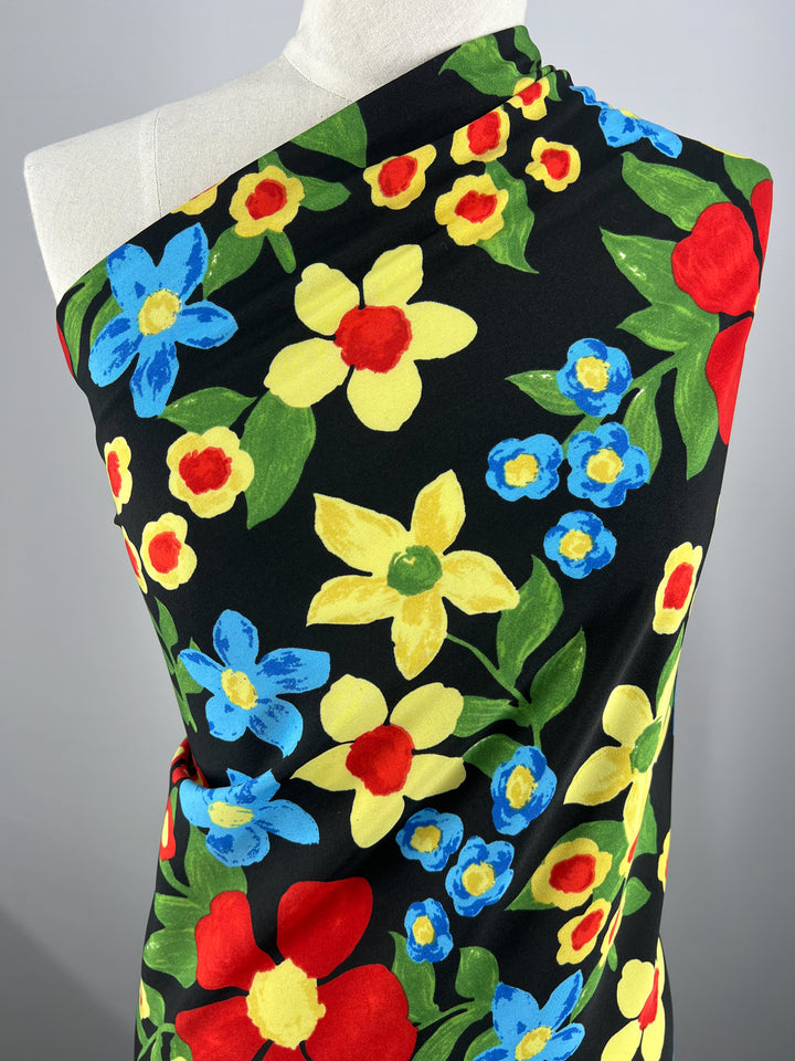 A designer fabric draped over a mannequin features a vibrant floral pattern with large red, blue, and yellow flowers on a black background. The colorful blossoms are surrounded by green leaves, creating a striking contrast against the dark backdrop. This is the Deluxe Print - Nursery - Black - 155cm by Super Cheap Fabrics.