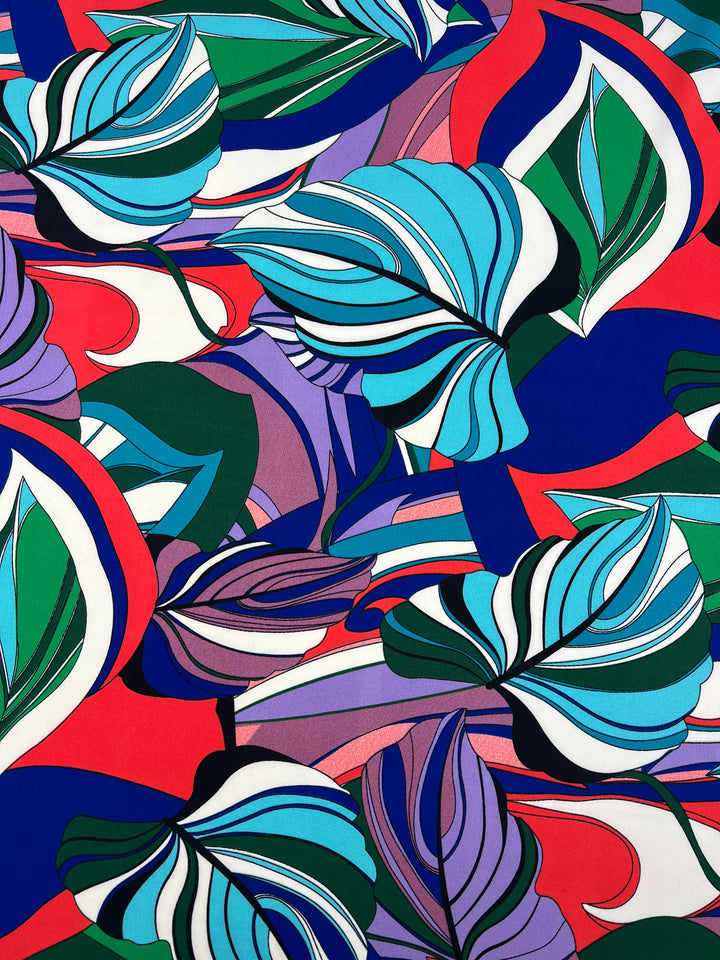 A vibrant, abstract pattern featuring large, swirling leaf-like shapes in various colors on a versatile polyester fabric. Predominant hues include teal, blue, green, red, purple, and white. The overlapping shapes create a dynamic, visually stimulating composition found in Super Cheap Fabrics' Deluxe Print - Jungle Tides - 155cm.