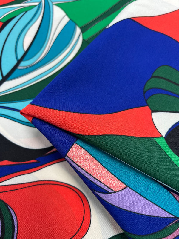 A colorful, folded piece of **Deluxe Print - Jungle Tides - 155cm by Super Cheap Fabrics** is displayed with a vibrant abstract pattern, featuring bold shapes in red, blue, green, and purple. The polyester fabric rests on top of another piece with a similarly vivid and intricate design.