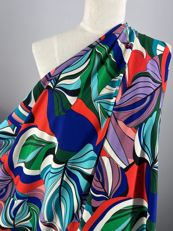 A mannequin draped with a vibrant, abstract-patterned designer fabric featuring bold swirls and shapes in red, blue, green, purple, and white. The intricate design showcases a lively and colorful combination of organic and geometric elements. This exquisite cloth is the Deluxe Print - Jungle Tides - 155cm from Super Cheap Fabrics.