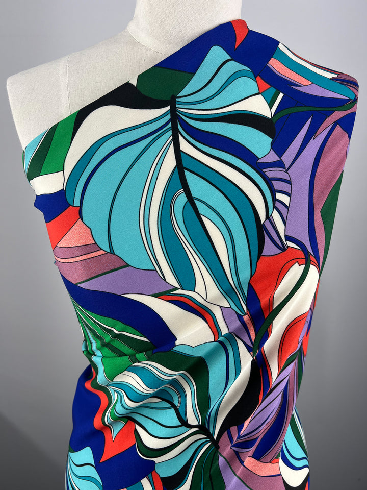 A mannequin is draped in a brightly colored, abstract-patterned **Super Cheap Fabrics Deluxe Print - Jungle Tides - 155cm** fabric featuring large, swirling leaf-like shapes in blue, green, white, purple, red, and black. The fabric has a smooth texture and the design is bold and eye-catching.