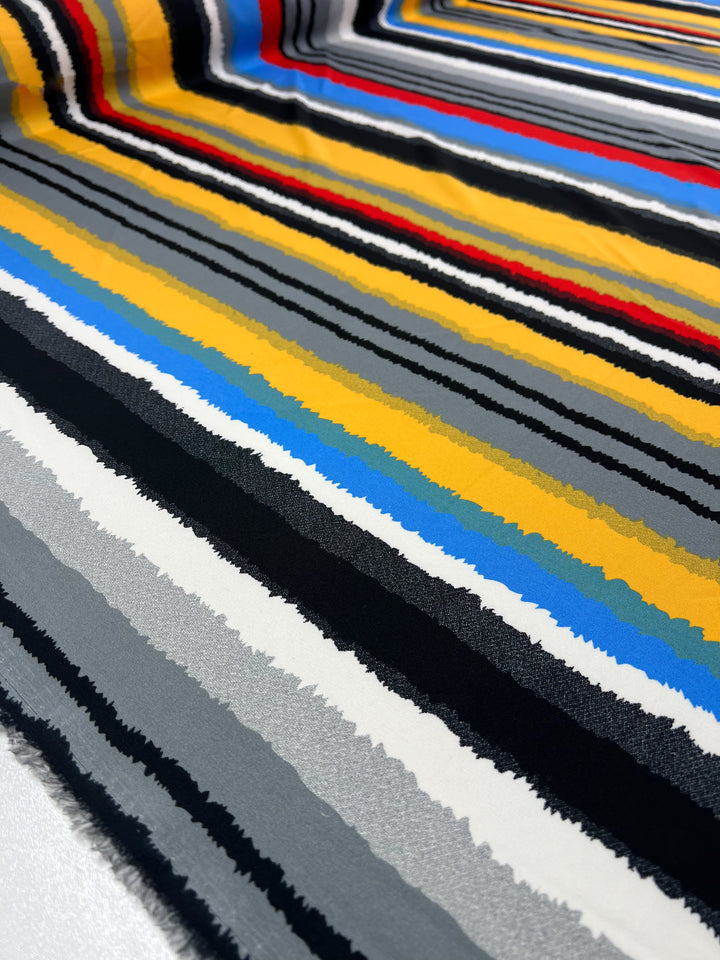 A fabric with a multicolored striped pattern is laid out. The stripes are horizontal with varying widths, featuring colors such as yellow, blue, red, black, gray, and white. This Deluxe Print - Neo - 155cm from Super Cheap Fabrics creates a visually striking, dynamic look.