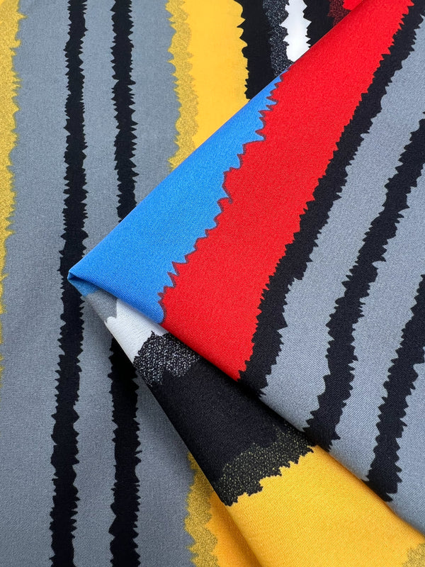 A close-up of a folded designer fabric featuring a bold, colorful pattern of jagged vertical stripes in black, grey, yellow, red, blue, and white creates a vibrant abstract design. This versatile polyester fabric from Super Cheap Fabrics is called Deluxe Print - Neo - 155cm and is perfect for making any project stand out.