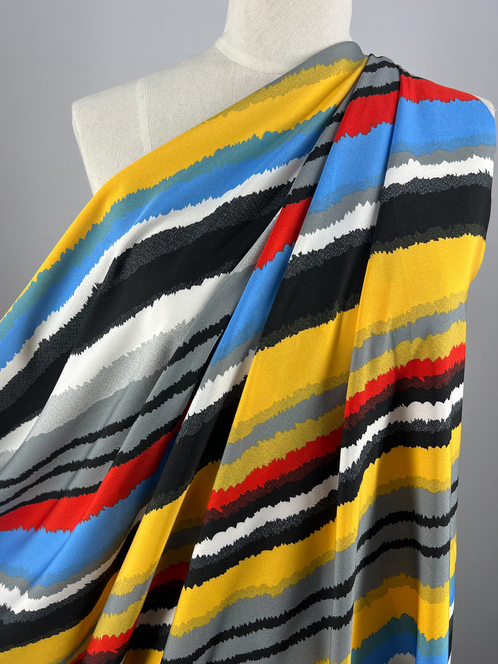 A close-up of a mannequin draped in vibrant polyester fabric with vivid horizontal stripes in various colors, including yellow, blue, black, red, gray, and white. This Deluxe Print - Neo - 155cm from Super Cheap Fabrics features a dynamic, wavy pattern that creates a striking visual effect.