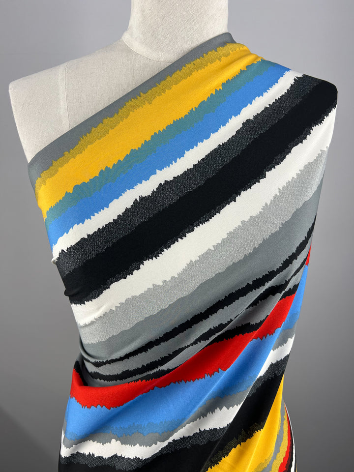 A mannequin is draped in a designer fabric featuring bold horizontal stripes in various colors, including yellow, blue, red, black, white, and shades of gray. The stripes have a textured, uneven edge creating a dynamic pattern on the versatile background of plain gray. This fabric is the Deluxe Print - Neo - 155cm by Super Cheap Fabrics.