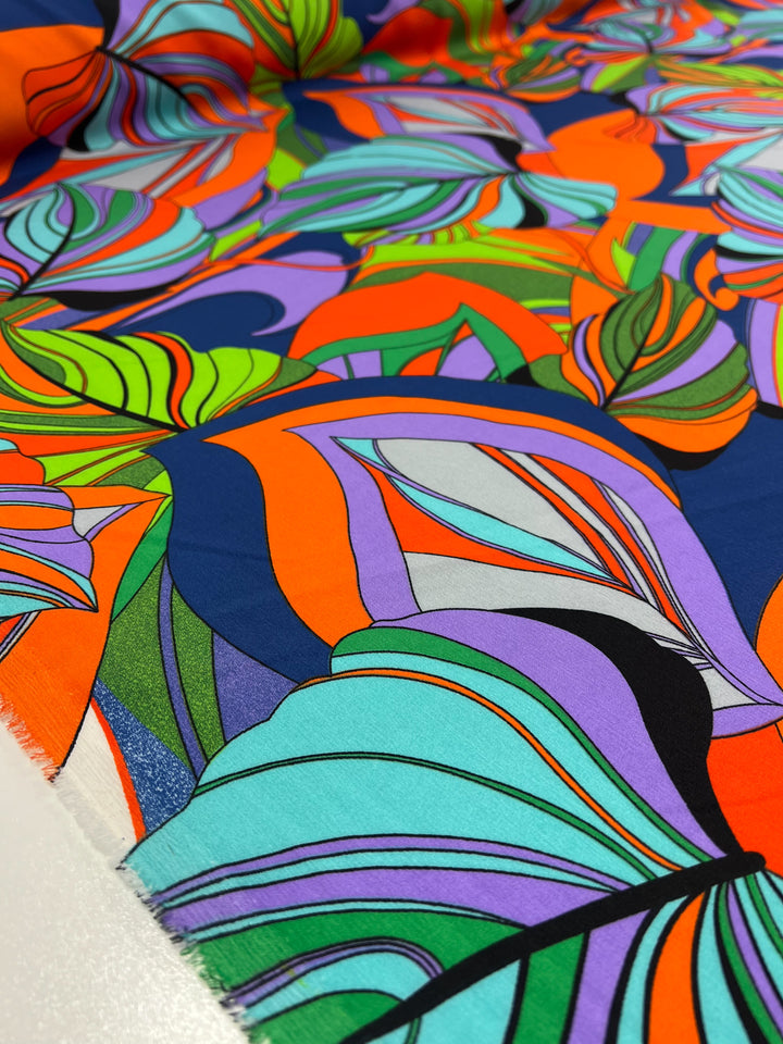 A close-up shot of a brightly colored polyester fabric featuring an abstract pattern. The design includes swirling shapes and overlapping leaves in bold hues of orange, blue, green, purple, and teal. The vibrant and dynamic composition of these bright prints creates a visually striking effect. Introducing the Deluxe Print - Terra - 155cm by Super Cheap Fabrics.