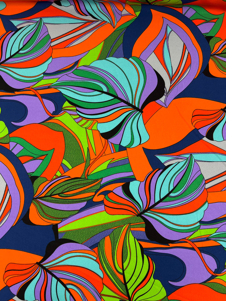 A vibrant polyester fabric pattern features an array of abstract, multi-colored leaves in shades of orange, green, purple, blue, and red. The bright prints are dynamic and rich, creating a sense of movement and energy against a dark background. Perfect for lovers of designer fabrics like the Deluxe Print - Terra - 155cm from Super Cheap Fabrics.