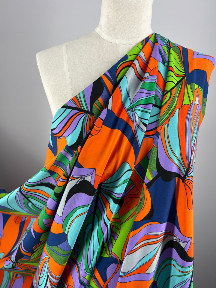 A mannequin draped in a vibrant, colorful polyester fabric featuring an abstract floral pattern in shades of orange, green, blue, purple, and black. The Super Cheap Fabrics Deluxe Print - Terra - 155cm designer fabric creates dynamic, flowing lines and a bold visual effect against the plain background.