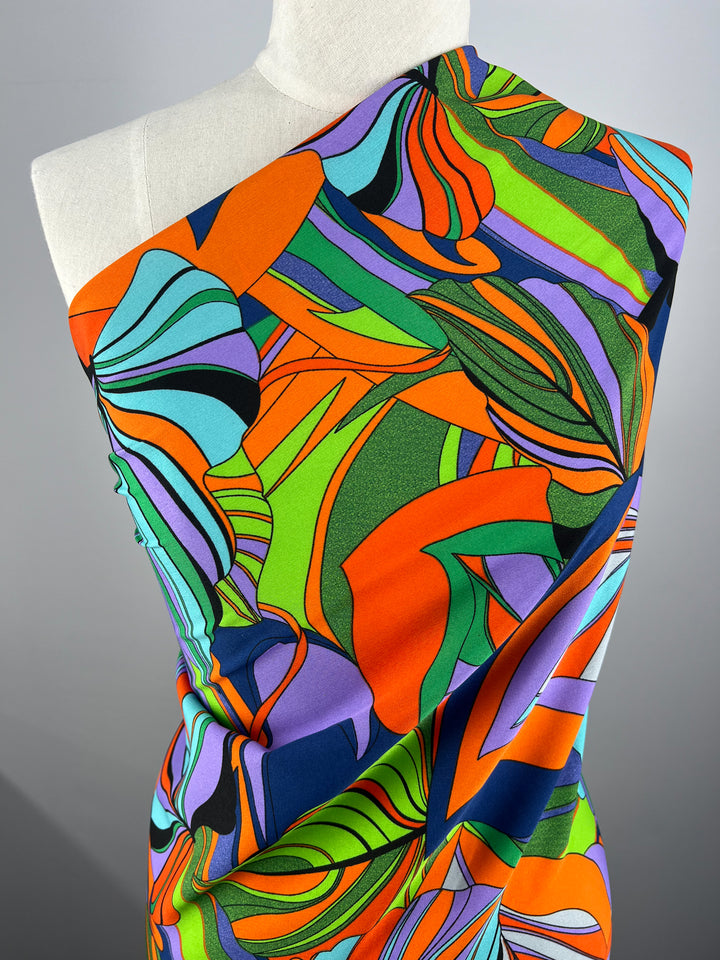 A mannequin is draped in vibrant, abstract-patterned polyester fabric featuring bold swirls and shapes in orange, green, blue, and purple. This Deluxe Print - Terra - 155cm by Super Cheap Fabrics boasts bright prints with a mix of curvy and angular forms, creating a dynamic and colorful visual effect.