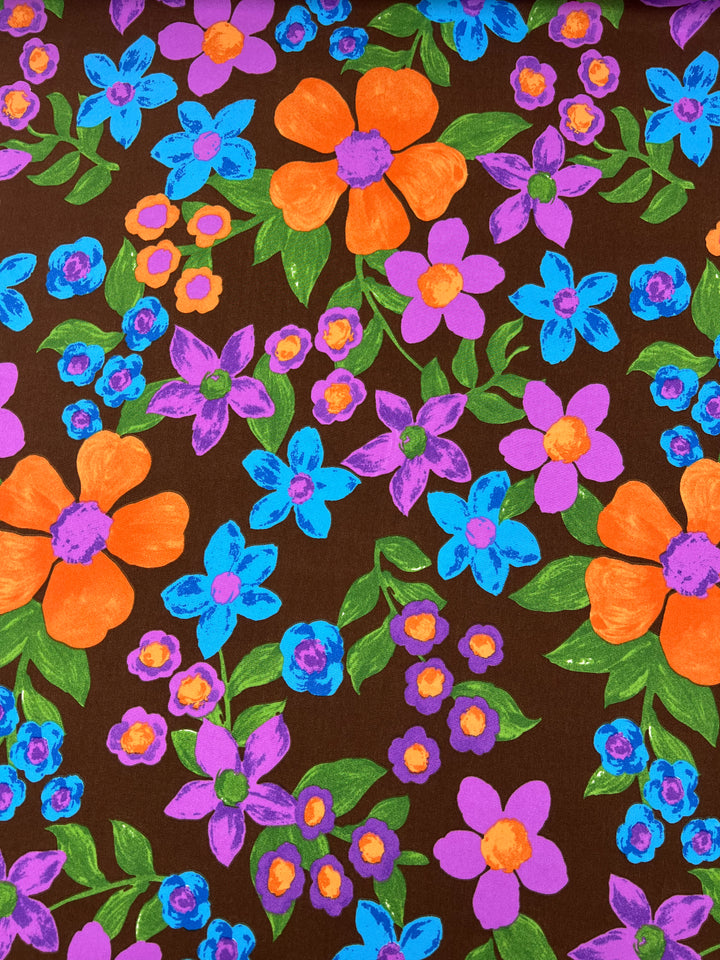 A vibrant designer fabric pattern featuring large orange flowers, medium-sized purple flowers, and small blue flowers with green leaves scattered across a dark brown background. The Deluxe Print - Nursery - Bison - 155cm by Super Cheap Fabrics adds a lively and cheerful touch, making it a versatile textile choice.