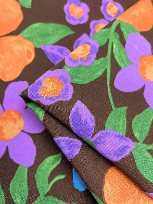 A close-up image of a brown polyester fabric with a vibrant floral print. The design features large orange and purple flowers with green leaves, showcasing the essence of Super Cheap Fabrics' Deluxe Print - Nursery - Bison - 155cm. A portion of the fabric is folded over, displaying more of the colorful pattern.
