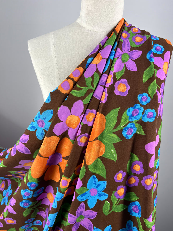 A mannequin draped in Super Cheap Fabrics' Deluxe Print - Nursery - Bison - 155cm, adorned with vibrant floral patterns, featuring large blue, purple, orange, and yellow flowers with green leaves. The designer fabric is elegantly arranged over one shoulder, showcasing the bold and colorful design.