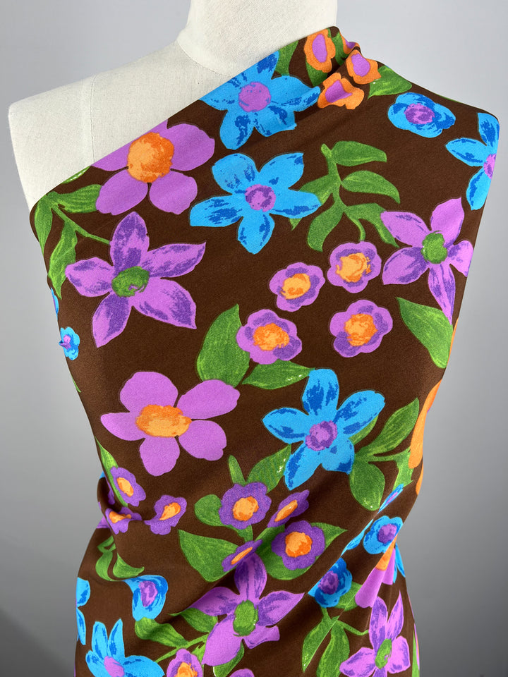 A close-up image of a mannequin draped in Super Cheap Fabrics' Deluxe Print - Nursery - Bison - 155cm. The versatile textile features large, vibrant flowers in shades of blue, purple, and orange against a brown background, with green leaves interspersed throughout.