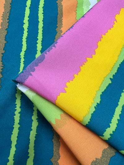 Close-up of colorful striped designer fabric. The design features vertical stripes in varying widths and colors, including blue, green, yellow, pink, orange, and white. The Deluxe Print - Lollipop - 155cm by Super Cheap Fabrics has a smooth polyester texture with a slight sheen, and the stripes have a painted, organic look.