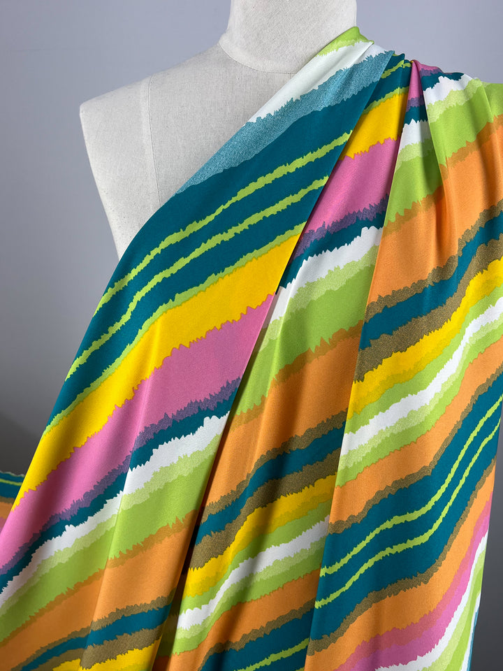 A mannequin draped with Super Cheap Fabrics' Deluxe Print - Lollipop - 155cm featuring horizontal, wavy, multicolored stripes. The stripes include shades of green, yellow, orange, pink, teal, and white, creating a bold and lively pattern against a plain background.