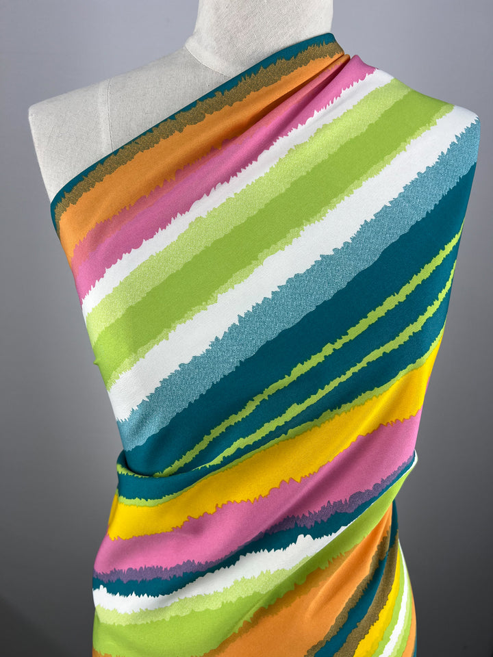 A mannequin draped in a vibrant, multi-colored polyester fabric featuring diagonal stripes in shades of teal, green, yellow, orange, pink, and white. The designer fabric **Deluxe Print - Lollipop - 155cm** by **Super Cheap Fabrics** has a textured appearance with each stripe blending smoothly into the next.
