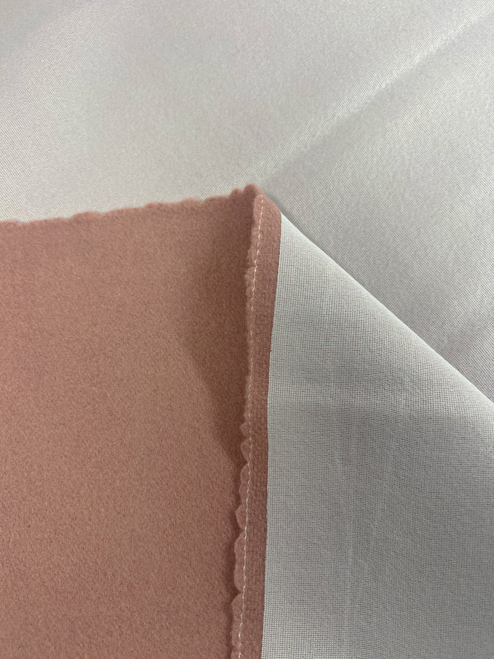 A close-up of a piece of Wool Cashmere - Cameo Brown - 150cm with a rough, scalloped edge from Super Cheap Fabrics. The main fabric is a light gray color, while the overlapping corner piece is a soft, dusty pink. Sustainably sourced and limited in stock, the textures and details are highlighted under the lighting.