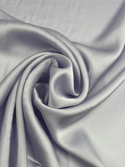 An elegant swirl of smooth, light grey Designer Viscose Satin - Lilac - 145cm from Super Cheap Fabrics is displayed in close-up. The fabric reflects light softly, accentuating its glossy texture and fluid drape. The folds create a sense of movement and luxurious softness, ideal for garment lining fabric.