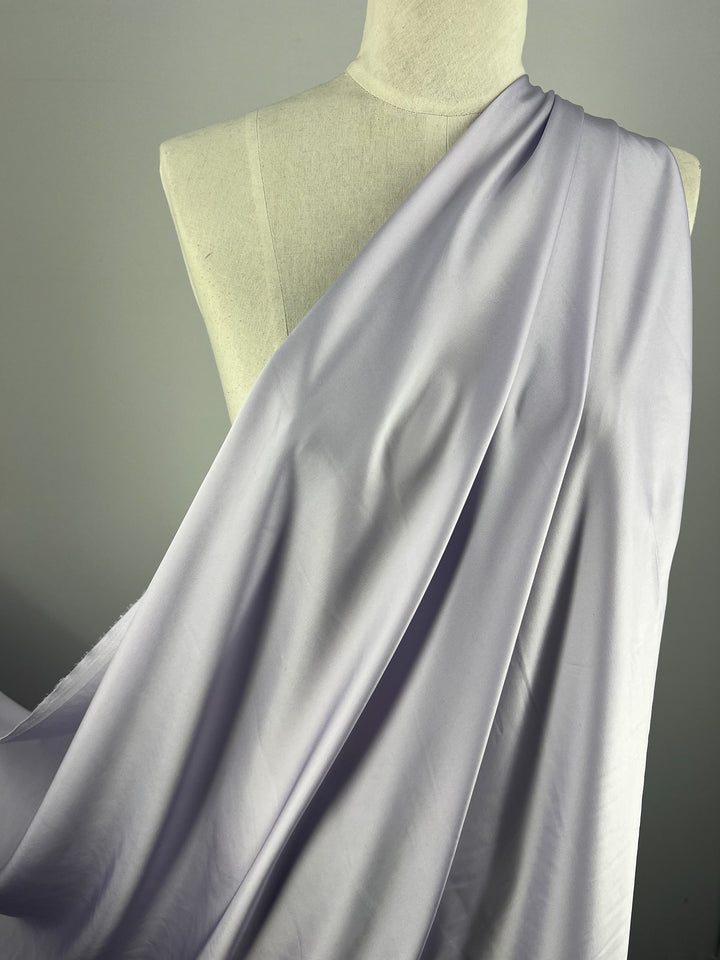 A mannequin draped with Super Cheap Fabrics' Designer Viscose Satin - Lilac - 145cm over one shoulder. The reflective sheen and subtle folds create a luxurious appearance. The background is a plain, light grey.