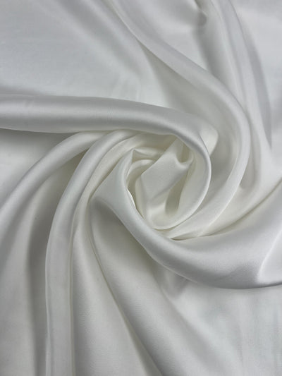 A close-up photo of ivory fabric, showcasing its smooth, glossy texture and elegant draping. The Designer Viscose Satin - Ivory - 140cm by Super Cheap Fabrics is arranged in soft, flowing folds with subtle highlights and shadows, emphasizing its luxurious and silky quality.