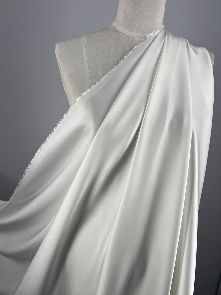 A white mannequin displays a draped, silky ivory fabric. The material flows smoothly over the mannequin's shoulder, showcasing a slight sheen and a delicate texture. This Designer Viscose Satin - Ivory - 140cm from Super Cheap Fabrics stands out against the plain gray background, emphasizing its elegance and exuding ex-designer haul vibes.