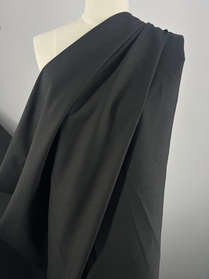 A versatile black fabric, possibly Cotton Suiting - Black - 150cm by Super Cheap Fabrics, is elegantly draped over a white dress form, forming smooth, flowing folds. The image captures the subtle texture and intricate layering of the material against a plain grey background.