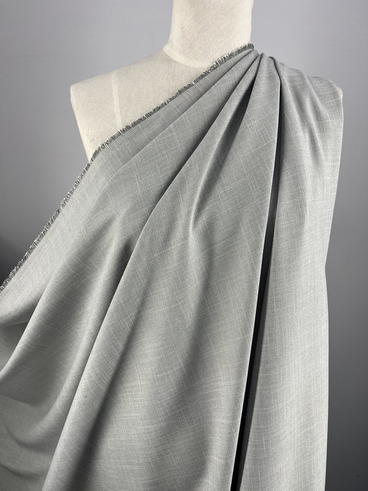 A close-up image of a mannequin draped with Super Cheap Fabrics' Textured Cotton Suiting - Grey - 150cm. The material has a fine, subtle texture and is neatly laid over the shoulder of the mannequin, emphasizing its smooth and flowing quality. Perfect for formal suits, this versatile fabric stands out against the neutral gray background.