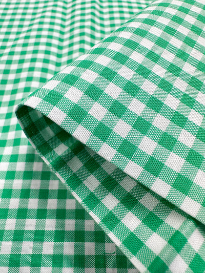 Close-up of the Cotton Gingham Mini - Green - 112cm from Super Cheap Fabrics, a folded piece of lightweight cotton fabric featuring a green and white checkered pattern. This fabric, crafted from natural cotton fibres, is displayed diagonally to highlight its texture and repeating design.