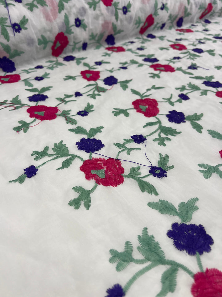 A close-up of a lightweight cotton fabric adorned with embroidered floral patterns in bright pink and deep purple, accompanied by green leaves. The intricate design lies flat on the surface, with the fabric roll partially visible in the background. This is the Embroidered Cotton - Pinkle Vines - 145cm from Super Cheap Fabrics.