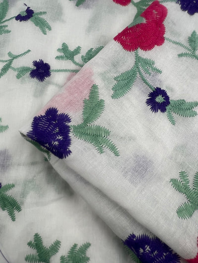 Close-up of Embroidered Cotton - Pinkle Vines - 145cm from Super Cheap Fabrics with green and purple floral designs. The detailed stitching forms intricate patterns, adding texture and color. The lightweight fabric is partially folded, enhancing its soft and delicate appearance.