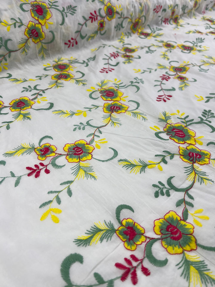 A close-up of Embroidered Cotton - MonaSun - 145cm by Super Cheap Fabrics adorned with intricate floral embroidery. The design features vibrant yellow, red, and green flowers connected by green stems and leaves, creating a delicate and colorful pattern. The fabric's texture is elegantly visible through the embroidery.