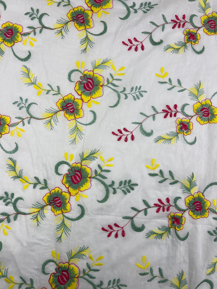 A white lightweight embroidered cotton fabric featuring a floral pattern with yellow and red flowers, surrounded by green leaves and stems. The flowers are scattered evenly across the 100% cotton fabric, creating a symmetrical and vibrant design. This is the Embroidered Cotton - MonaSun - 145cm from Super Cheap Fabrics.