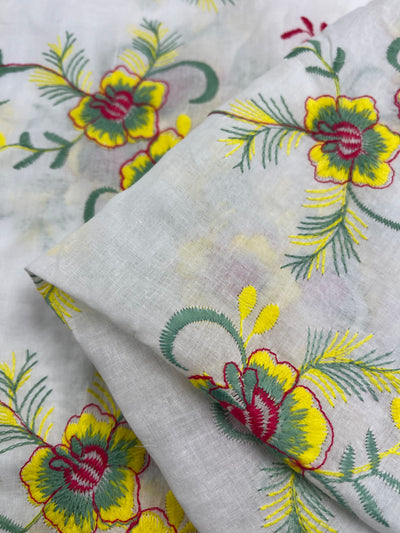 Close-up of lightweight embroidered cotton fabric showcasing a multi-colour floral design. The intricate embroidery includes yellow and green flowers with red accents, plus green leaves and stems. Some of the fabric is folded over to highlight the detailed, vibrant pattern. This is Embroidered Cotton - MonaSun - 145cm by Super Cheap Fabrics.