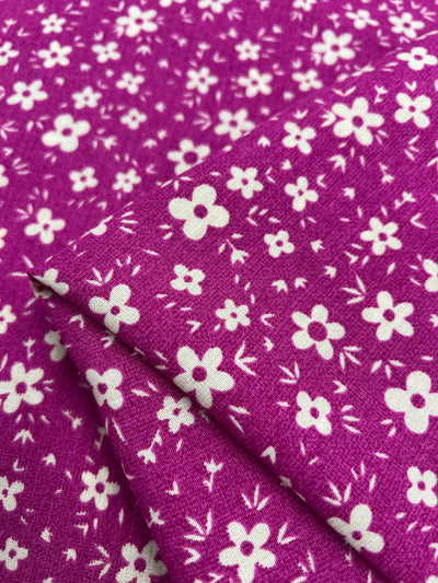 A close-up image of a beginner-friendly fabric with a purple background and a pattern of small white flowers. The lightweight fabric is folded neatly, creating layers that showcase the floral design. The texture appears soft with a slight sheen, ideal for **Super Cheap Fabrics** Bamboo Rayon - Pink DaisyRain - 145cm enthusiasts.