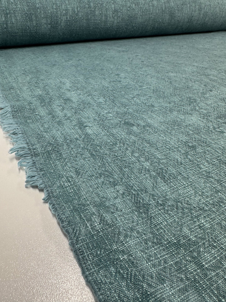 A close-up view of a roll of the Peacock-colored Basket Dyed Cotton from Super Cheap Fabrics reveals its soft, slightly textured surface with fringed edges on the left. This 100% cotton upholstery fabric, measuring 140cm wide, showcases a subtle basket weave that enhances its woven appearance.