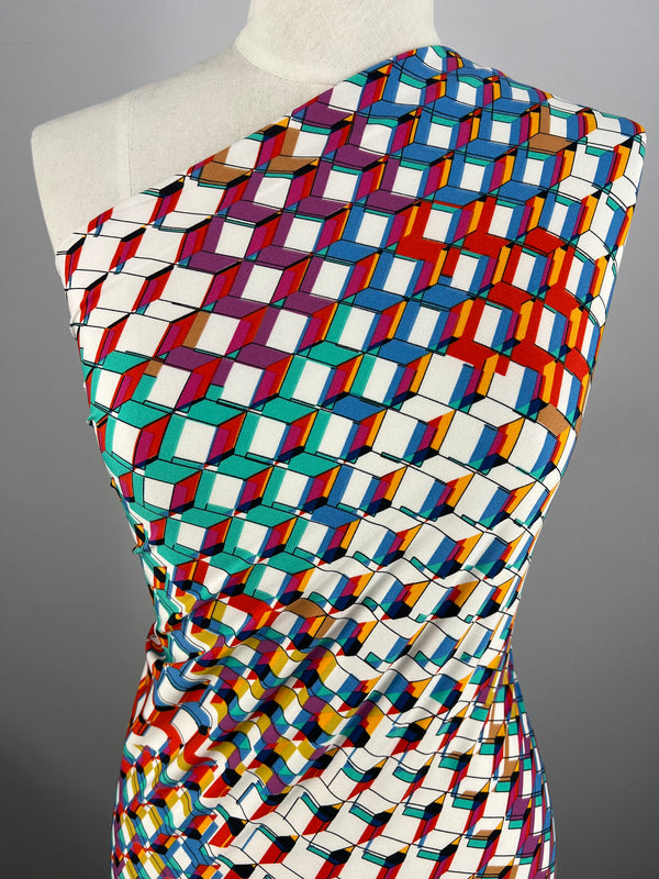 A close-up of a mannequin wearing a one-shoulder dress made from "Printed Lycra - Rubex - 150cm" by Super Cheap Fabrics, featuring a vibrant geometric print. The pattern consists of colorful interconnected cubes in hues of red, blue, green, and purple, creating an optical illusion effect against a plain gray background.