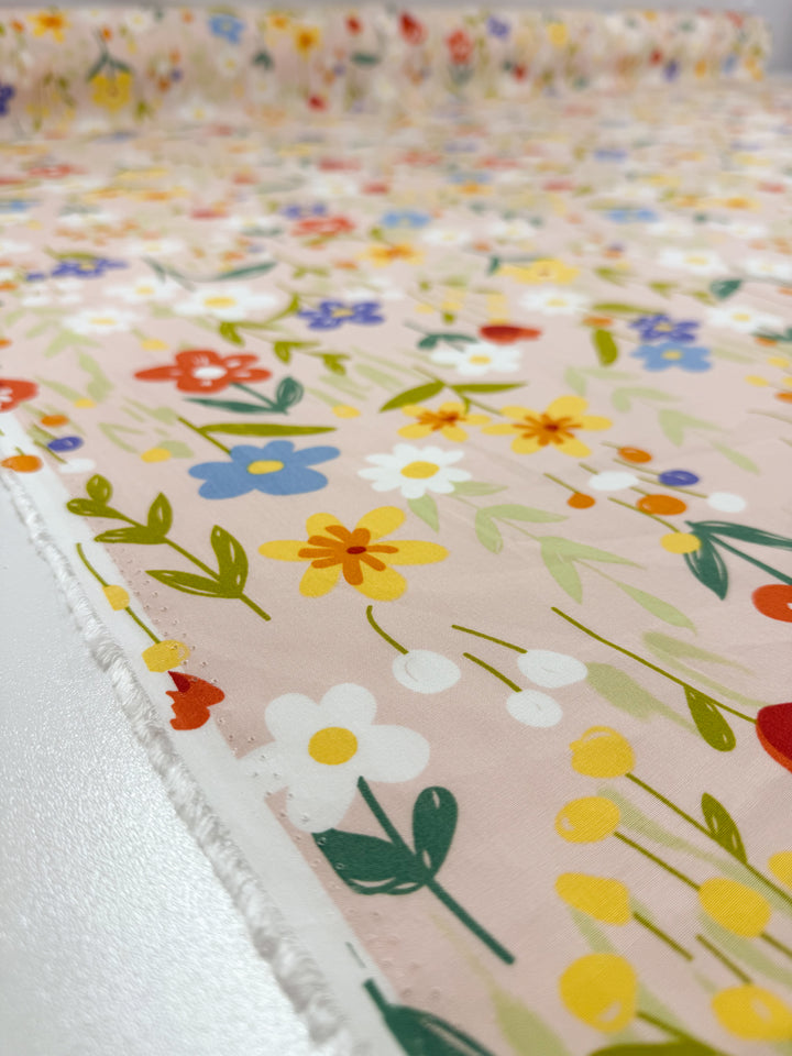 A close-up of Super Cheap Fabrics' Designer Rayon - Meadow Mingle (145cm) shows vibrant yellow, white, blue, and orange floral prints on pink, with a roll of this versatile fabric in the background.