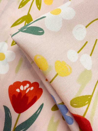 A close-up of Super Cheap Fabrics’ Designer Rayon - Meadow Mingle (145cm), showcasing light pink fabric with red, white, and yellow floral prints, green leaves, and folded edges—perfect for versatile projects.
