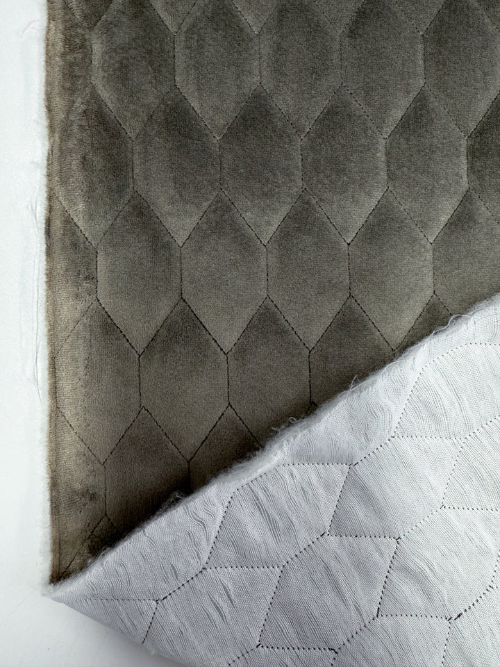 A close-up of Super Cheap Fabrics’ Quilted Velvet - Vintage Khaki (145cm) shows its smooth, grayish-brown geometric hexagon pattern on top; the white underside with visible stitching is partially folded to reveal both sides.