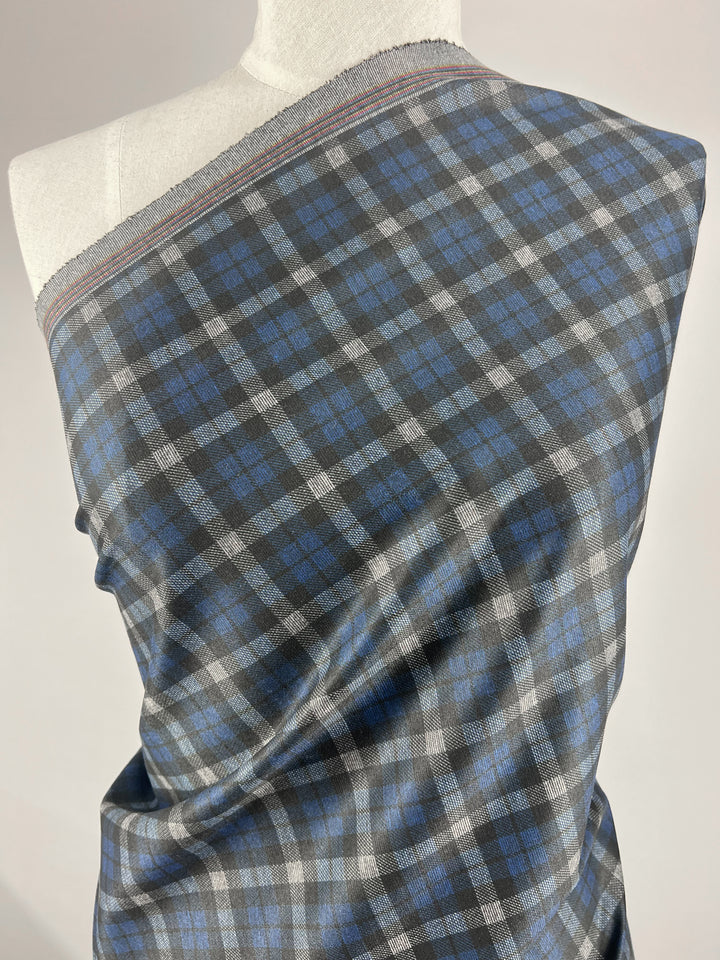 A mannequin is draped with a piece of Super Cheap Fabrics' Ponte - Checkers fabric, measuring 150cm and exhibiting a medium-weight structure in grey, blue, and black plaid. The checkered pattern features alternating blue and black squares intersected by thin grey lines, making it an ideal choice for pants. The edges of this polyester fabric are adorned with a red and white stitch pattern.