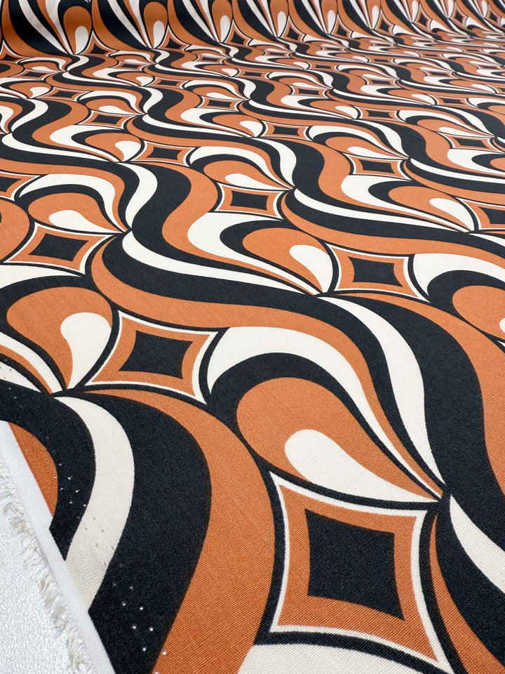 A close-up of Super Cheap Fabrics’ Designer Rayon - Retro Swirl (142cm), showcasing retro wavy geometric black, orange, and cream patterns with repeating diamond and oval motifs.