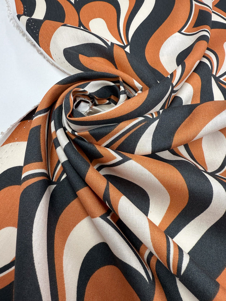 A close-up of Super Cheap Fabrics' Designer Rayon - Retro Swirl (142cm), featuring a retro wavy geometric pattern in black, brown, and cream, with fabric edges visible in the background.