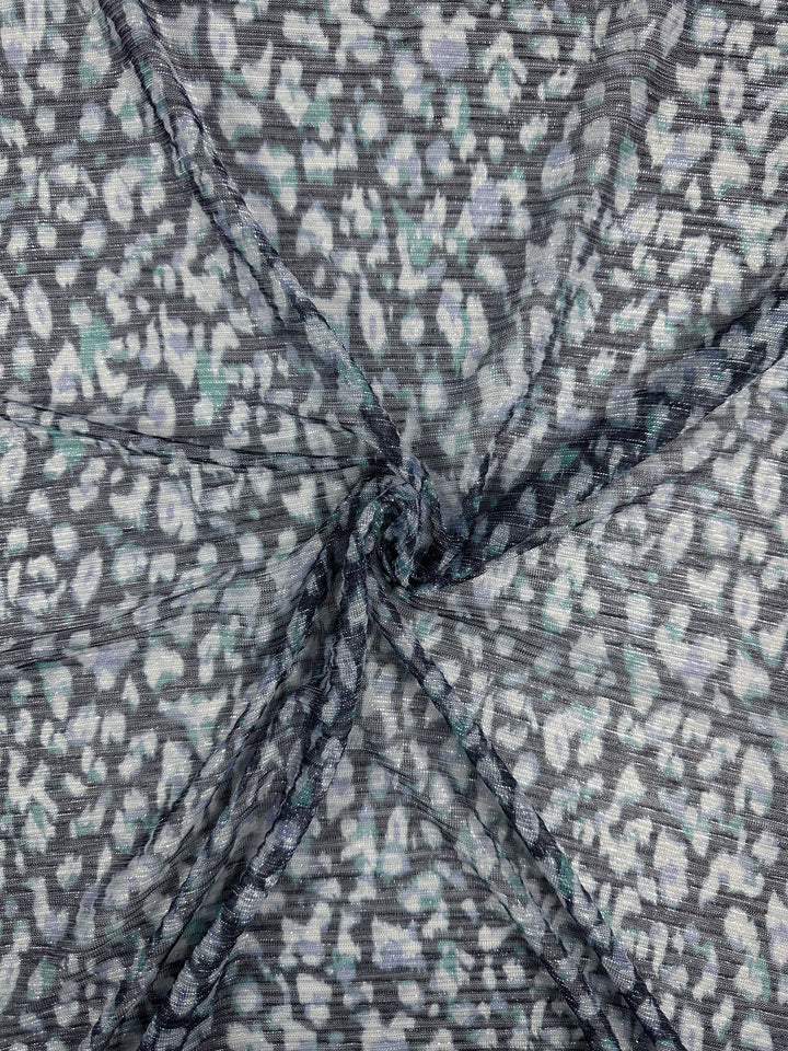 Close-up of Metallix Pleat - Ocean Leopard fabric by Super Cheap Fabrics, showcasing its textured, semi-transparent polyester with a twisted center. This gray and navy material displays a subtle animal print pattern reminiscent of leopard spots, blending shades of white and light gray for a soft effect. The fabric measures 140cm in width.